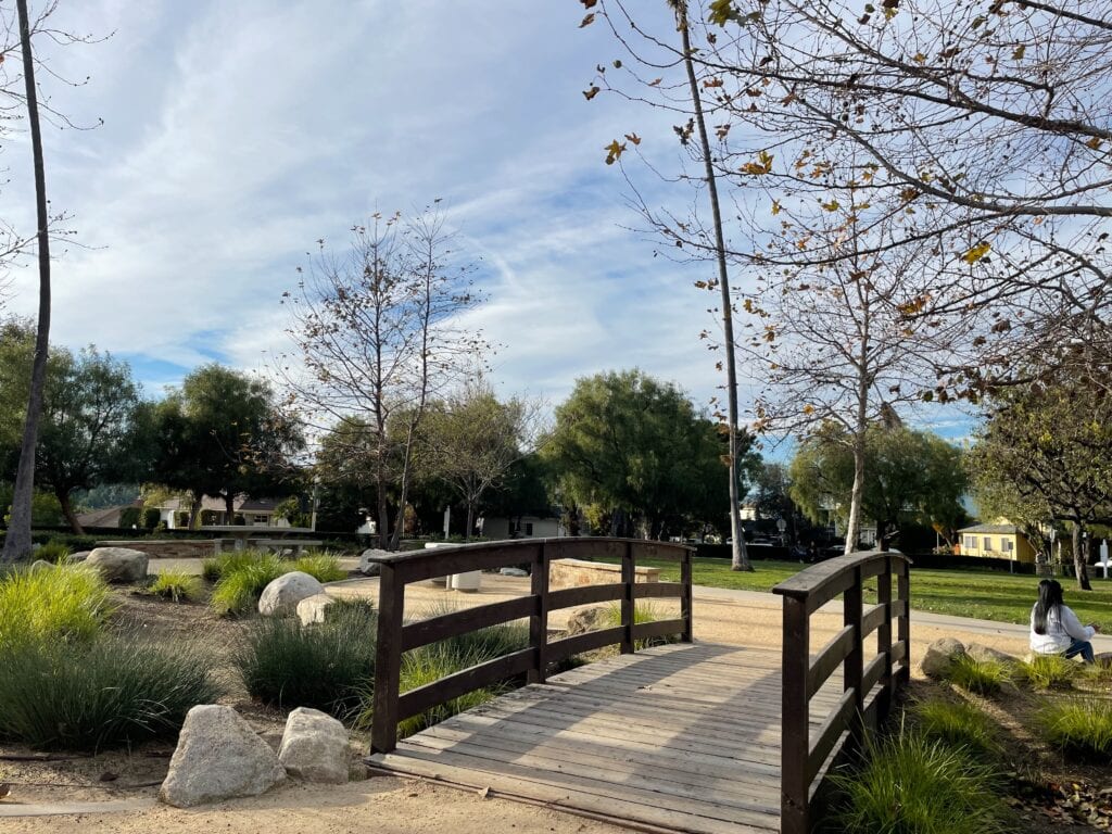 A brown mini bridge in the middle of circle park with large decorative boulders surrounding it.