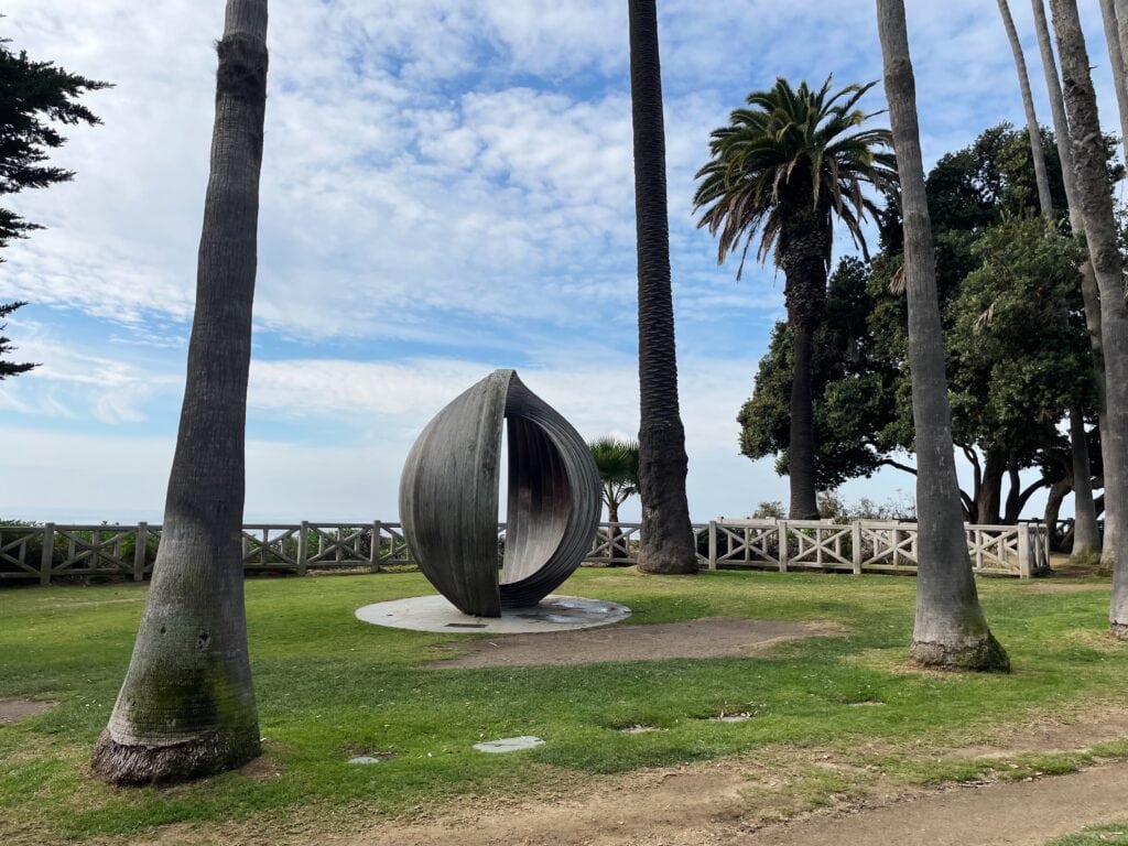 A grey oval shaped sculpture placed i the middle of a grassy patch overlooking the Pacific ocean.