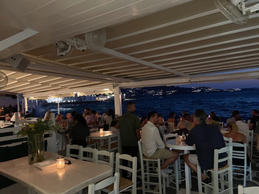 Diners eating dinner while watching the waves crash and the sun go down in Mykonos, Greece.